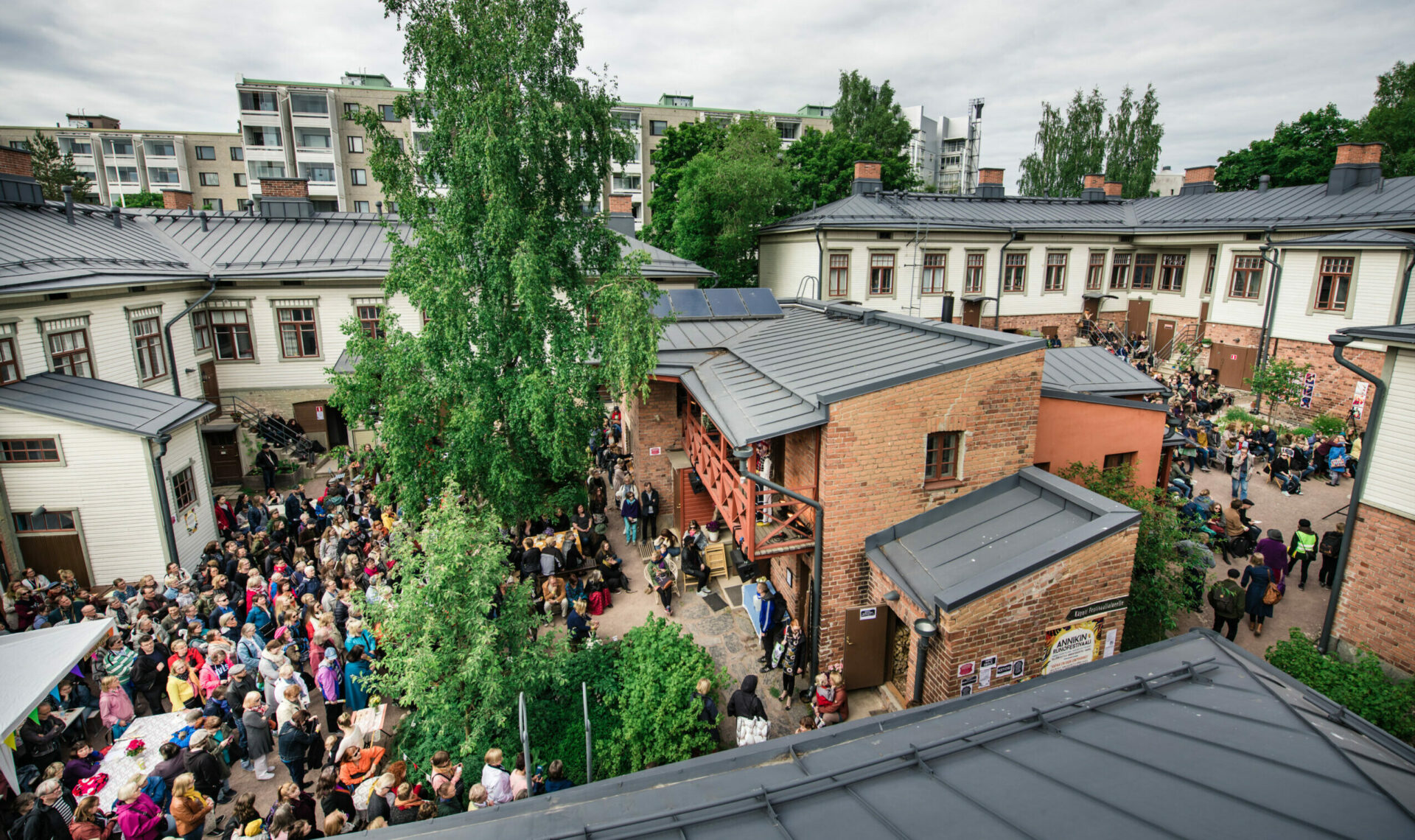 Annikin puutalokorttelin sisäpiha kuvattuna ylhäältä. Festivaalin yleisö katsoo esityksiä pihan kahdella puolella, keskellä on tiilinen rakennus ja iso vihreä koivupuu.