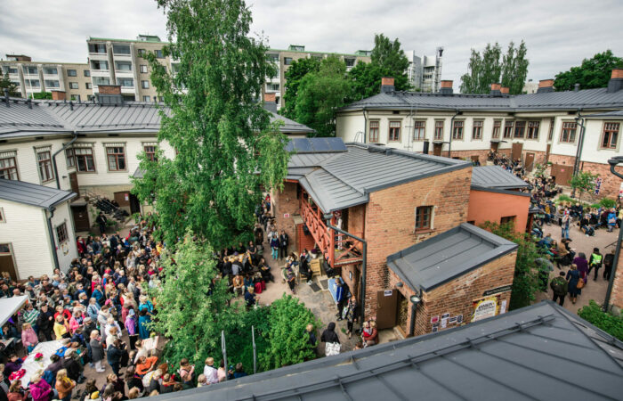 Annikin puutalokorttelin sisäpiha kuvattuna ylhäältä. Festivaalin yleisö katsoo esityksiä pihan kahdella puolella, keskellä on tiilinen rakennus ja iso vihreä koivupuu.