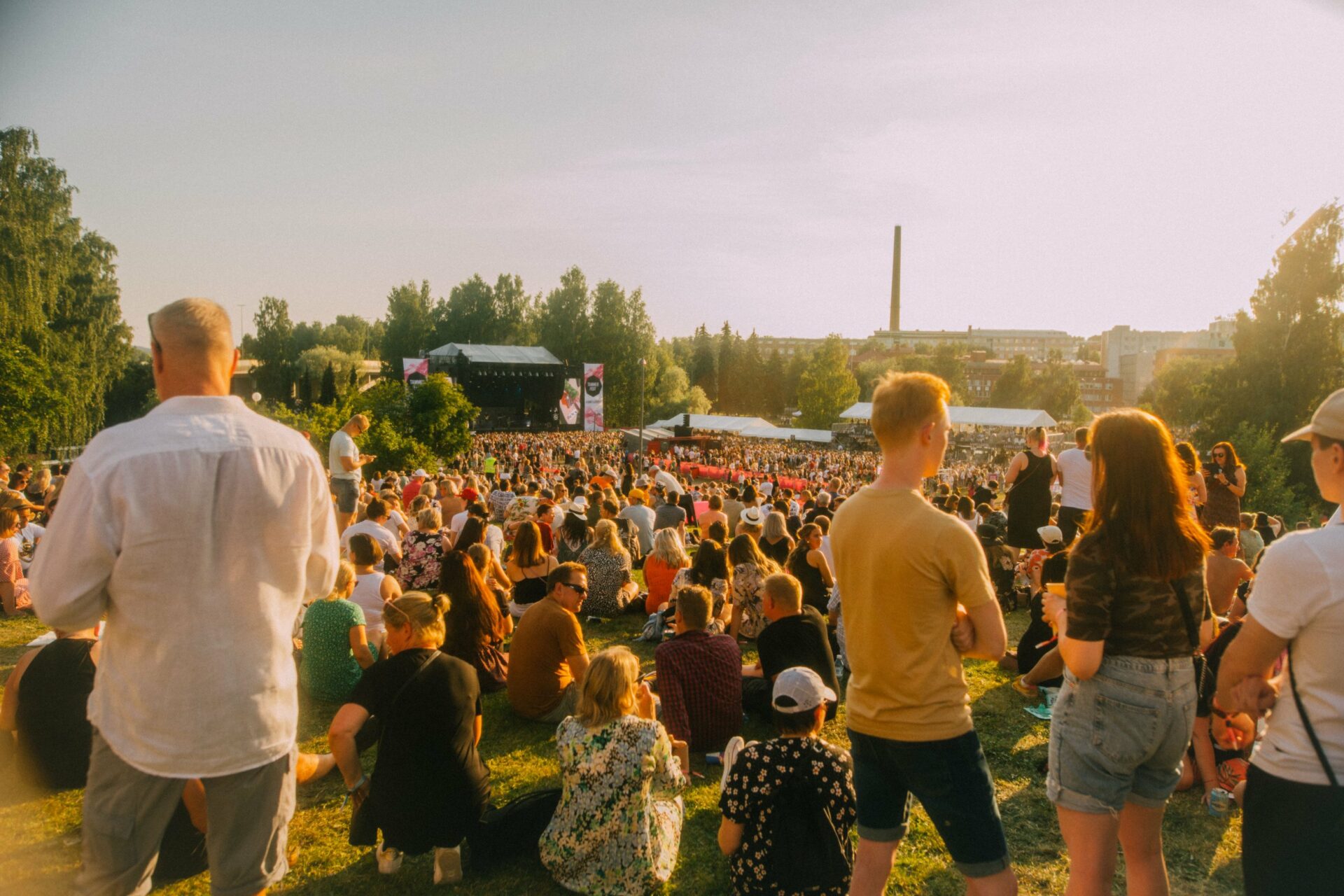 Yleisöä Tammerfestissa 2022 aurinkoisena päivänä. Osa yleisöstä istuu nurmikolla, taka-alalla näkyy Tammerfestin päälava, ja alue on vehreiden puiden ympäröimä.