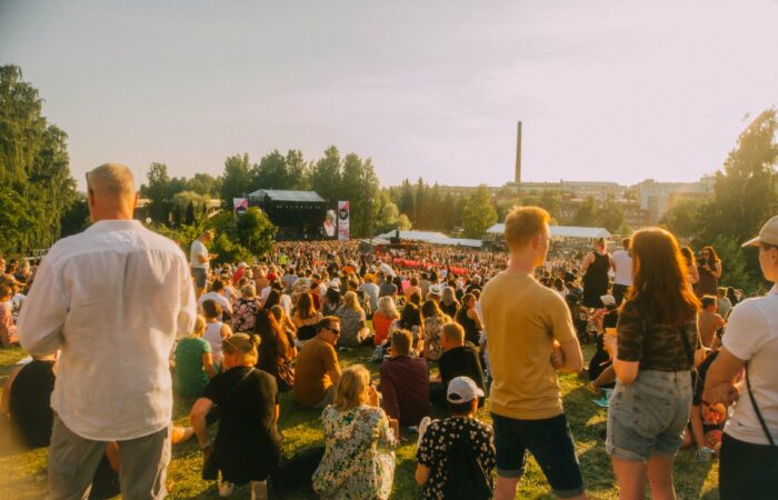 Yleisöä Tammerfestissa 2022 aurinkoisena päivänä. Osa yleisöstä istuu nurmikolla, taka-alalla näkyy Tammerfestin päälava, ja alue on vehreiden puiden ympäröimä.