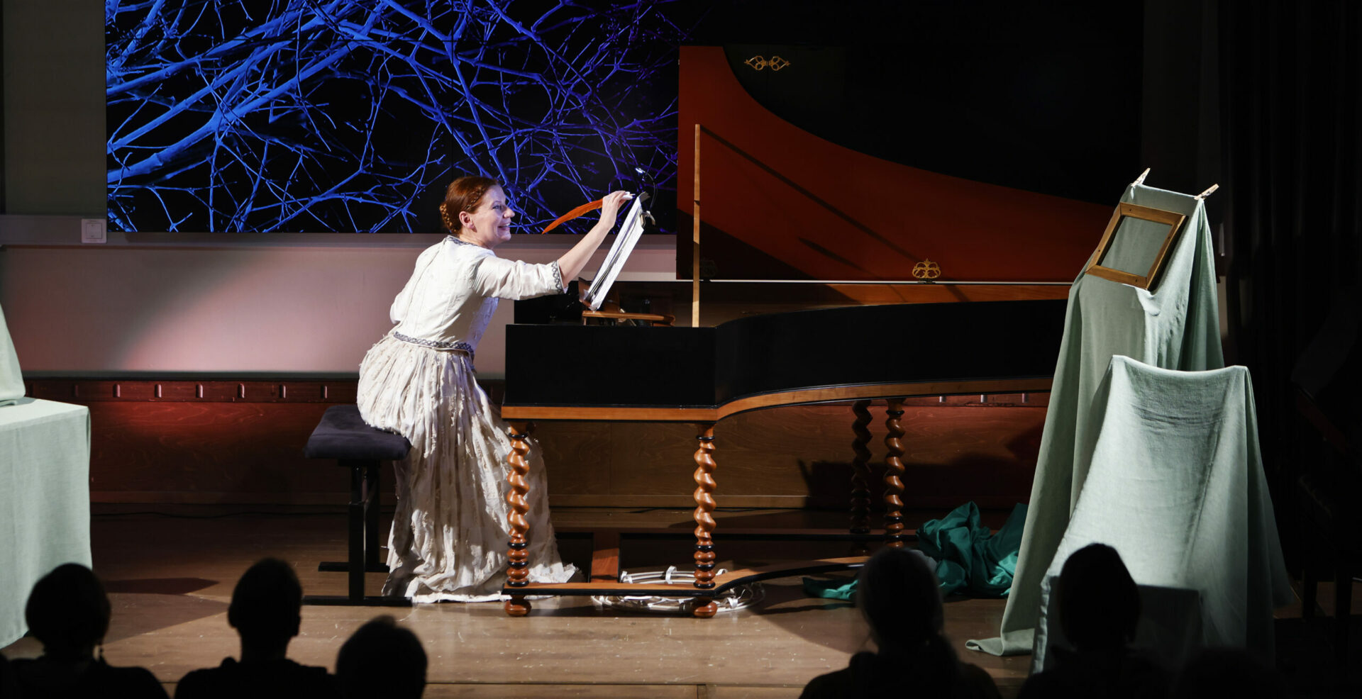 Laura Maddalena esityksen näyttelijä piirtää sulkakynällä nuottivihkoon flyygelin ääressä. Taustascreenillä sinisävyinen oksateos ja Tampere Biennale -teksti.