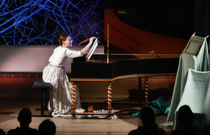 Laura Maddalena esityksen näyttelijä piirtää sulkakynällä nuottivihkoon flyygelin ääressä. Taustascreenillä sinisävyinen oksateos ja Tampere Biennale -teksti.