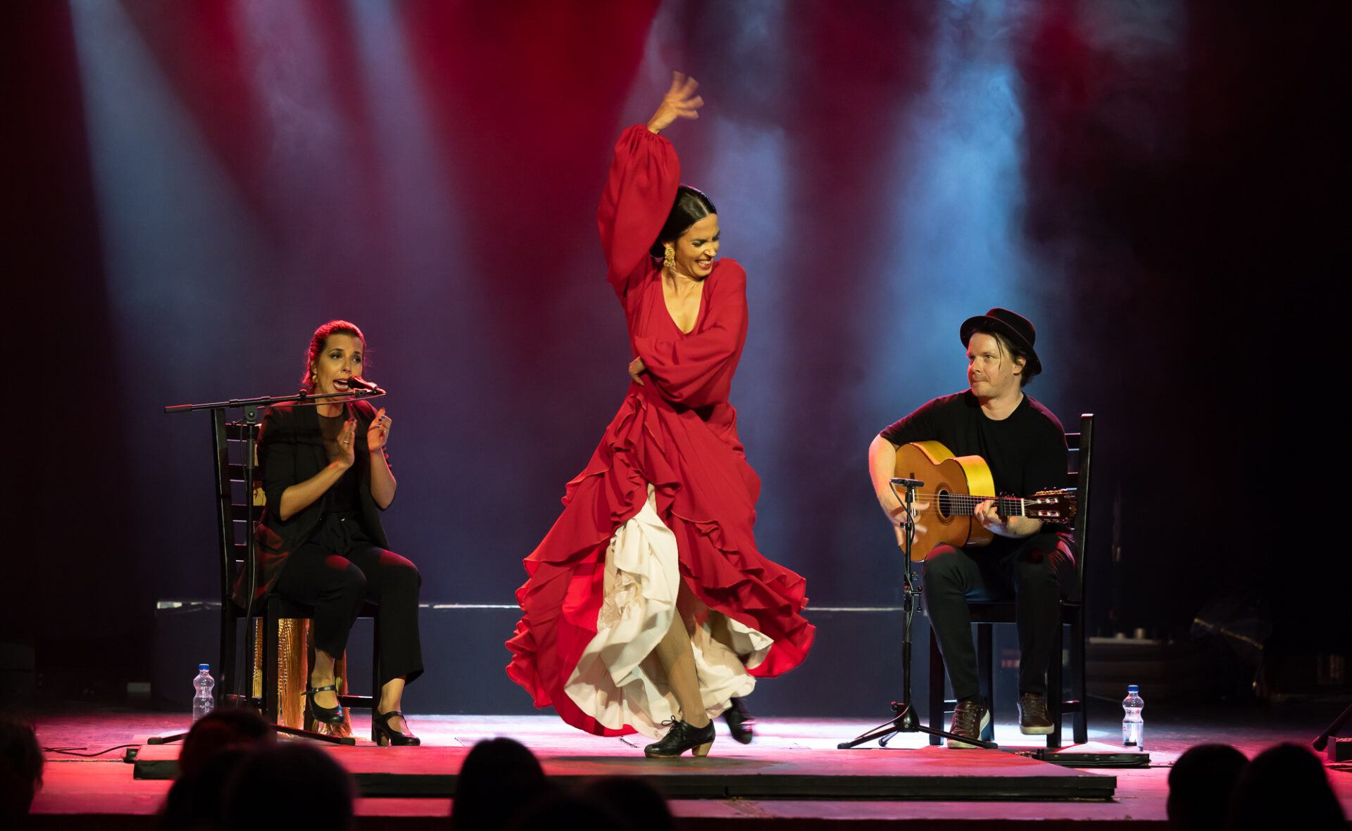 Brindamos Flamenco trio esiintymässä. Reunoilla kaksi istuvaa muusikkoa ja keskellä liikkeeseen eläytyvä tanssija punaisessa mekossa.
