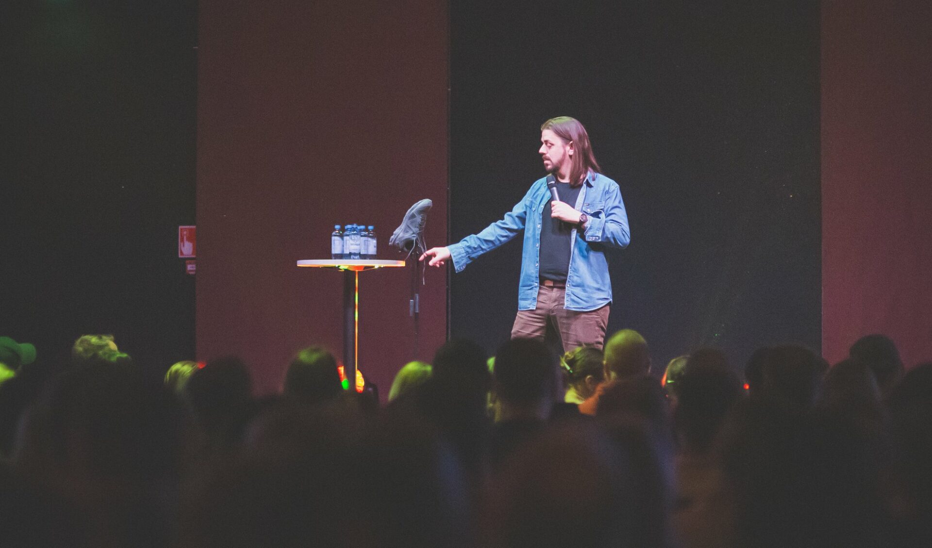 Lavalla koomikko Anders Helenius seisoo pystypöydän vieressä, pitää kädessään mikrofonia ja koskee mikkitelineeseen kiinnitettyä kenkää