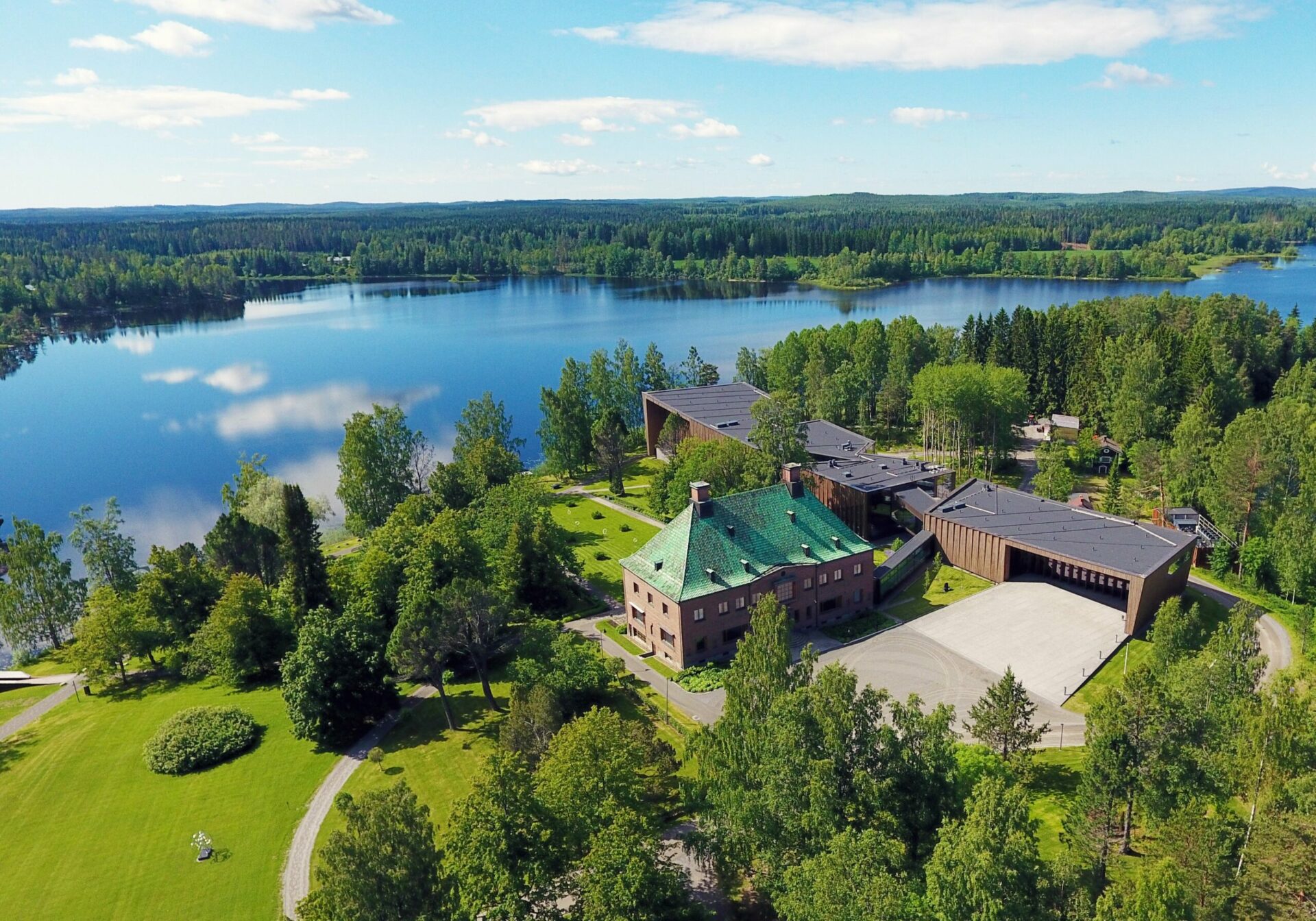 Ilmakuva Mäntän Serlachius-museo Göstasta. Aurinkoisessa maisemassa näkyvät rakennukset, vihreää nurmea, puita ja sinisenä heijastuva järvi.