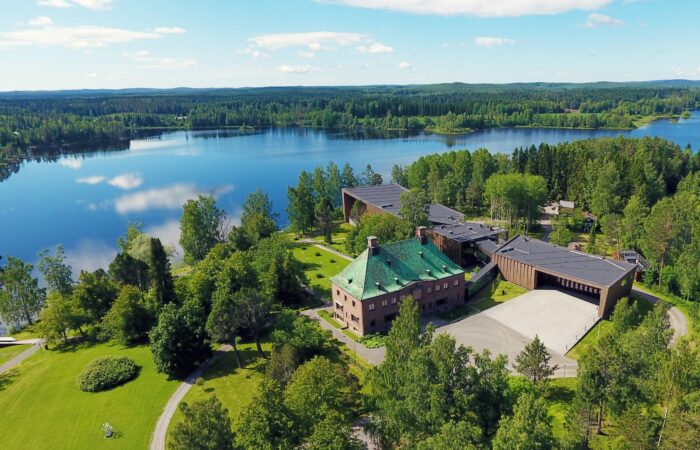 Ilmakuva Mäntän Serlachius-museo Göstasta. Aurinkoisessa maisemassa näkyvät rakennukset, vihreää nurmea, puita ja sinisenä heijastuva järvi.