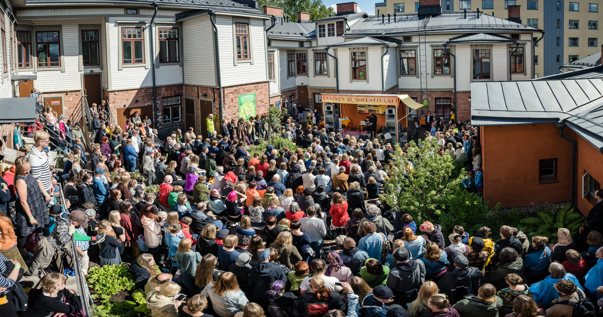 Runsaslukuinen yleisö katsoo aurinkoisena alkukesän päivänä esitystä Annikin runofestivaalilla. Yleisöä ympäröivät valko- ja vihreäsävyiset puutalot, kuvan oikeassa laidassa näkyy oranssiksi rapattu kivirakennus ja hiukan pihapensaita.