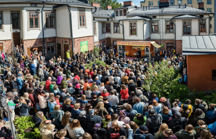 Runsaslukuinen yleisö katsoo aurinkoisena alkukesän päivänä esitystä Annikin runofestivaalilla. Yleisöä ympäröivät valko- ja vihreäsävyiset puutalot, kuvan oikeassa laidassa näkyy oranssiksi rapattu kivirakennus ja hiukan pihapensaita.