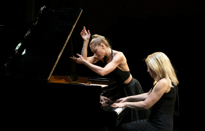 Mustan kuvan keskellä valaistuna balettitanssija Minna Tervamäki ja pianisti Heini Kärkkäinen flyygelin ääressä