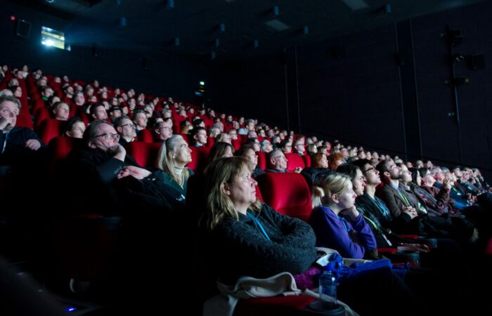 Yleisö täydessä ja pimeässä elokuvasalissa edestä päin kuvattuna valkokankaan valossa