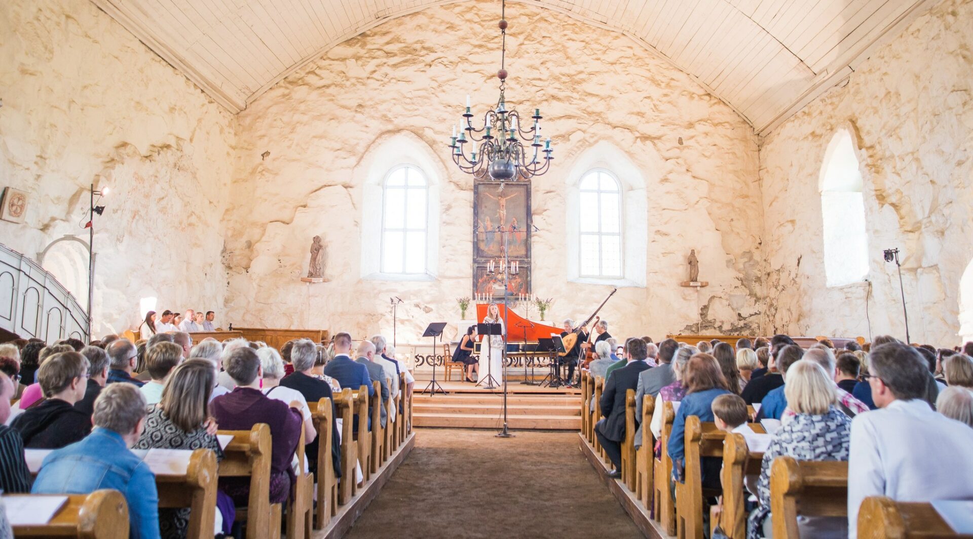 Konsertti vaaleaseinäisen kivikirkon sisällä, keskellä kuvaa on kattokruunu, esiintyjät ja kuvan reunoilla kirkon penkit ja yleisö takaa päin kuvattuna