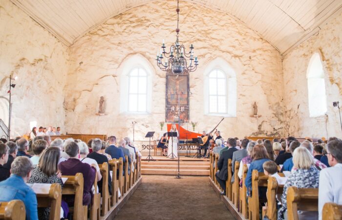 Konsertti vaaleaseinäisen kivikirkon sisällä, keskellä kuvaa on kattokruunu, esiintyjät ja kuvan reunoilla kirkon penkit ja yleisö takaa päin kuvattuna