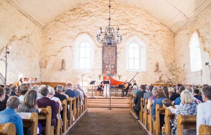 Konsertti vaaleaseinäisen kivikirkon sisällä, keskellä kuvaa on kattokruunu, esiintyjät ja kuvan reunoilla kirkon penkit ja yleisö takaa päin kuvattuna