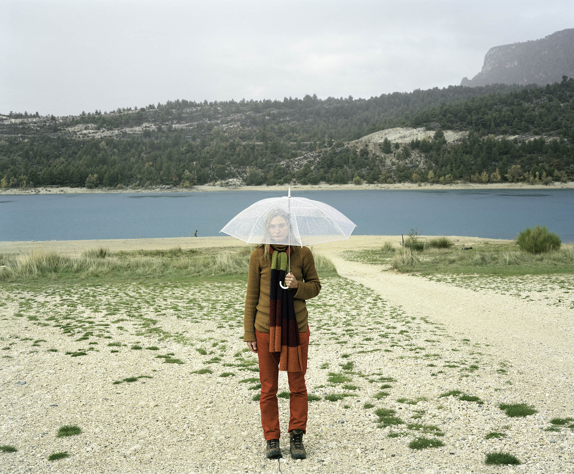 Henkilö seisoo rantahiekalla, kasvot läpinäkyvän sateenvarjon takana. Taustalla turkoosia vettä jonka takana havupuita ja kuiva hiekkarinne
