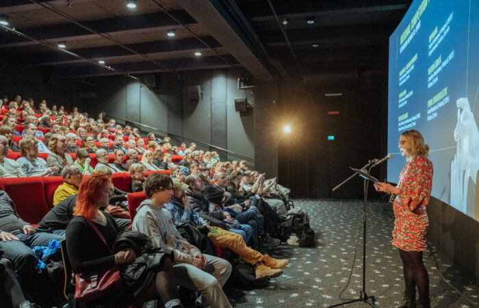 A host presenting a film screening to a full audience