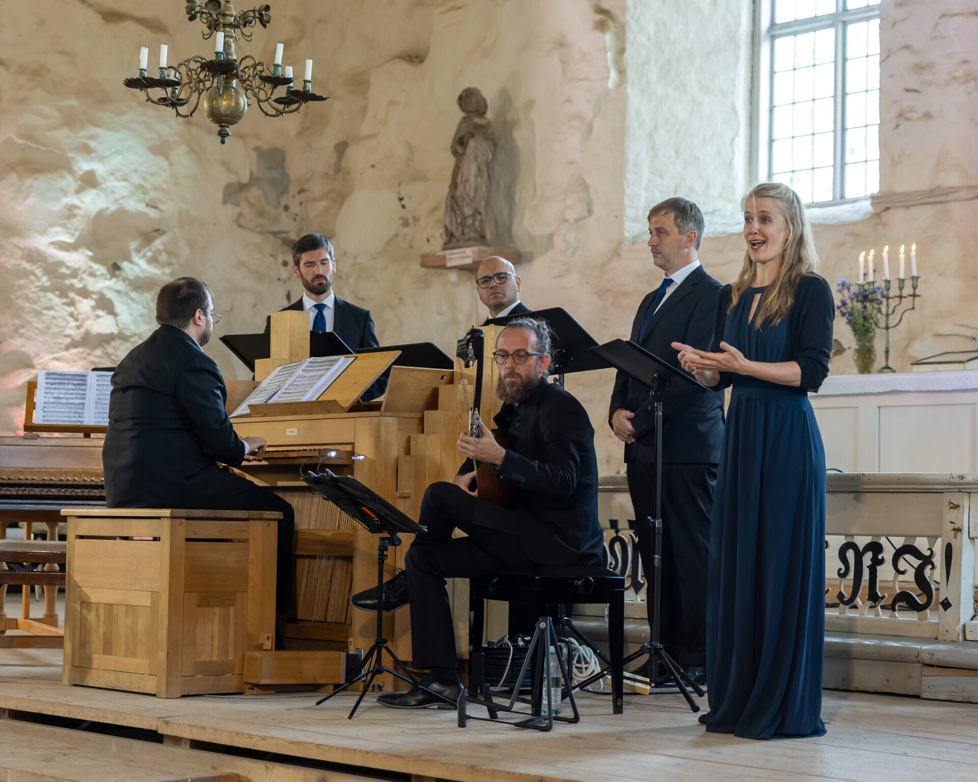 Venite Gentes esiintymässä Sastamala Gregorianassa 2024. Oikealla naishenkilö laulaa, keskellä mieshenkilö soittaa luuttua, vasemmalla mieshenkilö soittaa pianoa. Taustalla kolme muuta laulajaa.