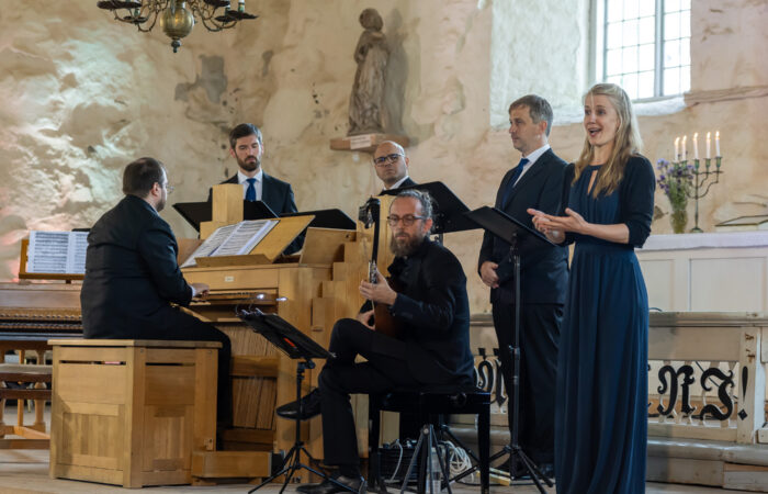 Venite Gentes esiintymässä Sastamala Gregorianassa 2024. Oikealla naishenkilö laulaa, keskellä mieshenkilö soittaa luuttua, vasemmalla mieshenkilö soittaa pianoa. Taustalla kolme muuta laulajaa.