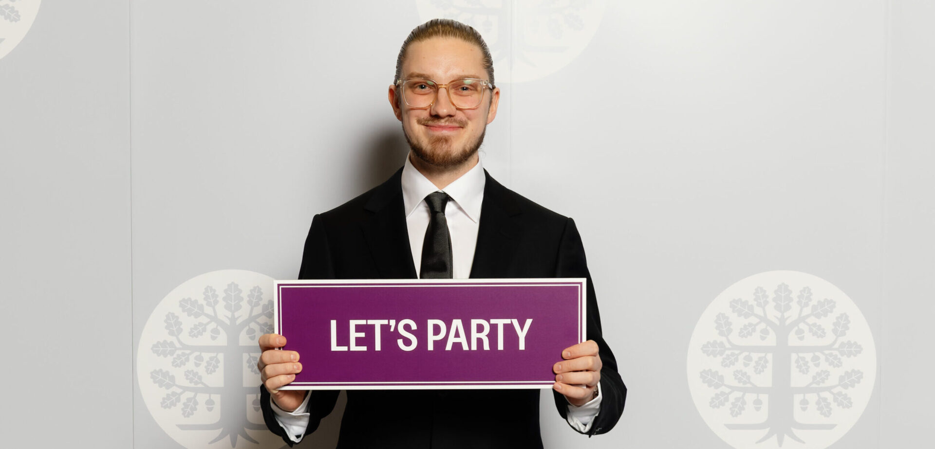 Johannes Rantanen valokuvausseinän edessä, kädessään violetti kyltti jossa lukee let's party.