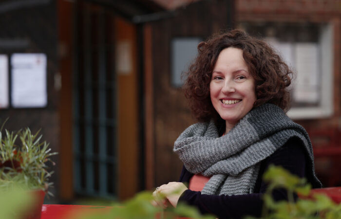 Hymyilevä Martyna Van Nieuwland istuu ulkona punaisen pöydän ääressä harmaa huivi päällään