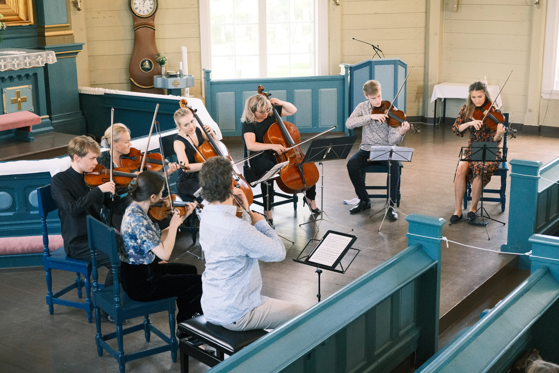 Kahdeksan muusikkoa esiintyy puisen kirkon alttarilla. Muusikoista kuusi soittaa viulua ja kaksi selloa.