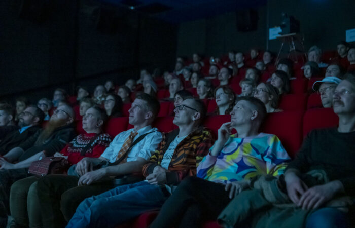 Kuva ihmisistä istumassa elokuvateatterin katsomossa. Kuva on otettu ihmisiä kohti. Teatterisalin valot ovat sammutettu.