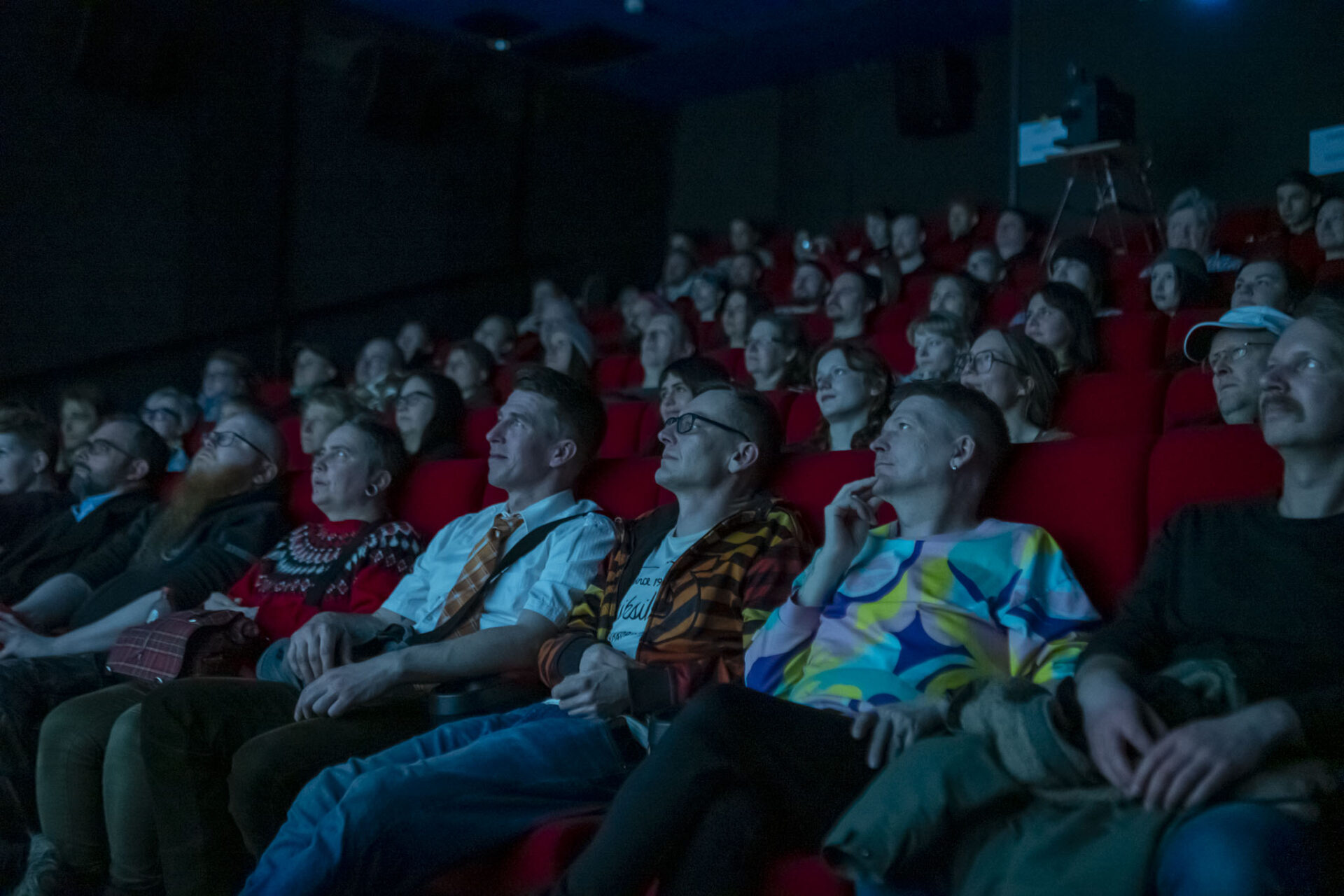 Kuva ihmisistä istumassa elokuvateatterin katsomossa. Kuva on otettu ihmisiä kohti. Teatterisalin valot ovat sammutettu.
