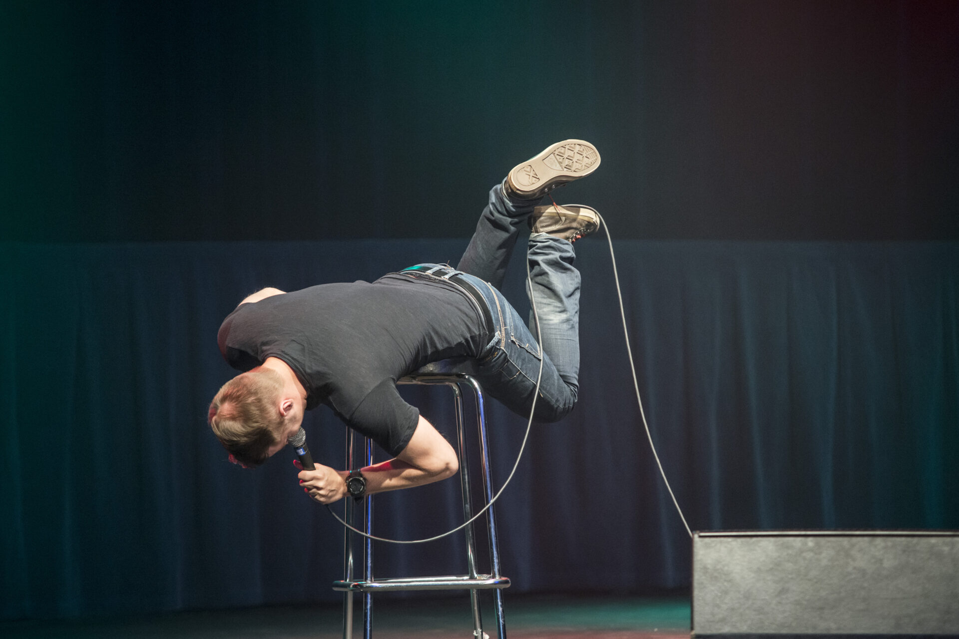 Stand up -koomikko lavalla. Koomikko on vatsallaan jakkaralla ja kädessä hänellä on mikrofoni.