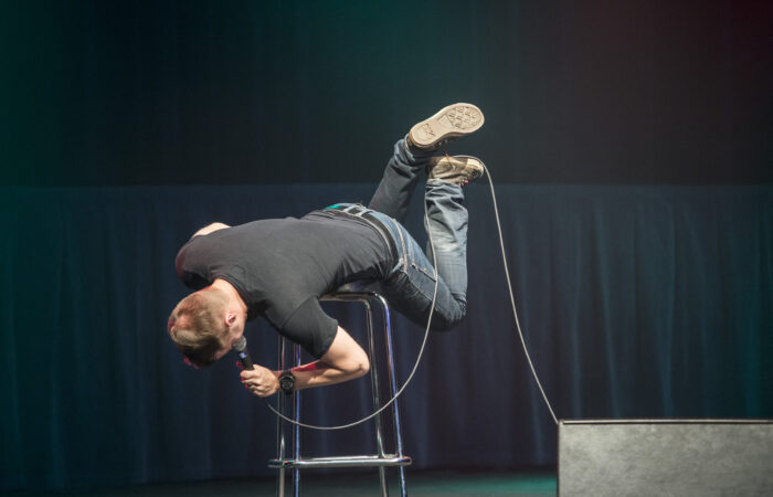 Stand up -koomikko lavalla. Koomikko on vatsallaan jakkaralla ja kädessä hänellä on mikrofoni.