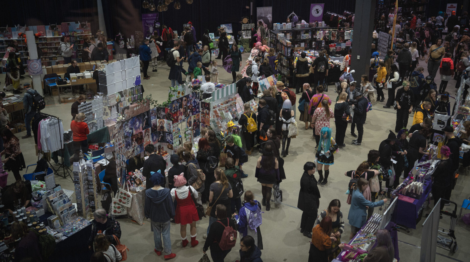 Tampere Kuplii tapahtuma Tampere-talossa. Sisätiloissa useita myyntipöytiä ja kojuja täynnä taidetta, printtejä, käsitöitä ja oheistuotteita. Paikalla on runsaasti ihmisiä kiertämässä ja selaamassa tuotteita, osa pukeutuneena värikkäisiin tai cosplay-tyylisiin asuihin.