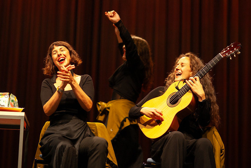 Laulaja ja kitaristi esiintyvät istuen. Taustalla musta-asuinen tanssija.