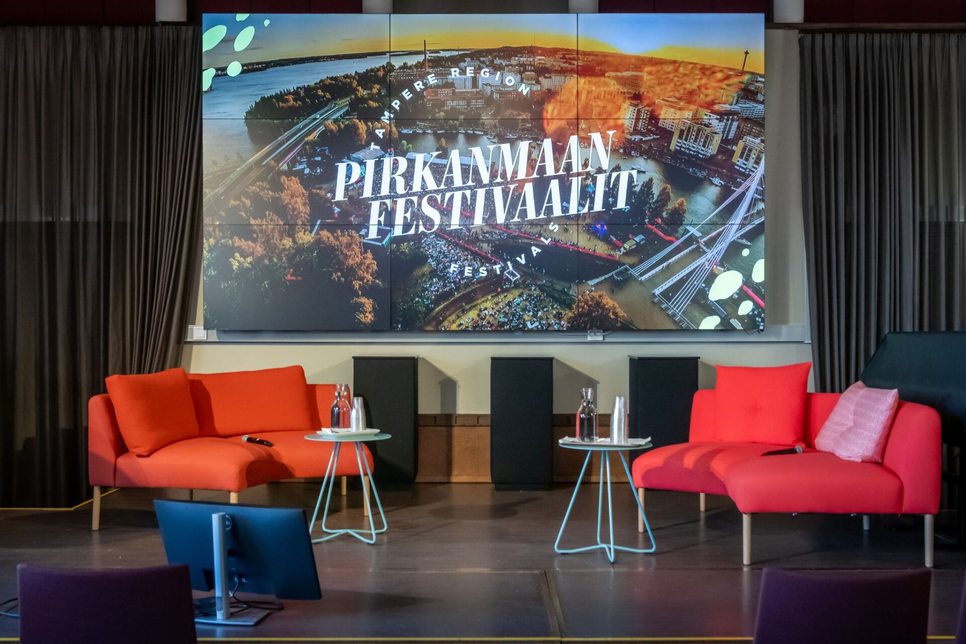 Kuvassa iso näyttö, jossa Pirkanmaan festivaalien logo. Näytön edustalla tuoleja.