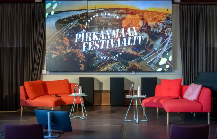 Kuvassa iso näyttö, jossa Pirkanmaan festivaalien logo. Näytön edustalla tuoleja.